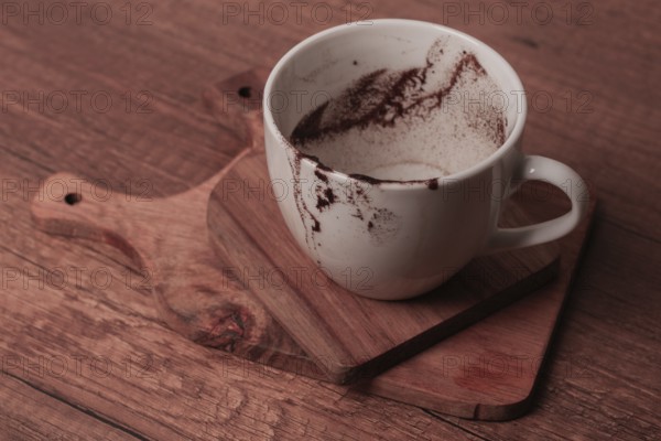 A white cup rests on a wooden cutting board showing remnants of coffee. The cozy setting suggests a recent enjoyment of a warm beverage, perfect for a relaxed moment