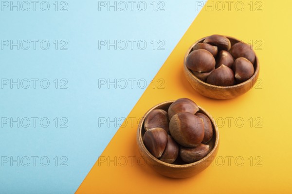 Wooden bowl with raw edible Ð¡hestnuts on blue and orange paper pastel background, side view, copy space, minimalism