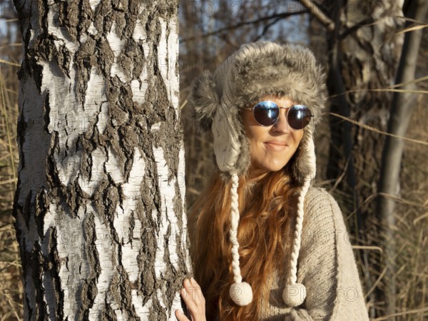 Slender pretty woman with fur hat and pompoms, red hair and warm winter colors between birch forest and reeds