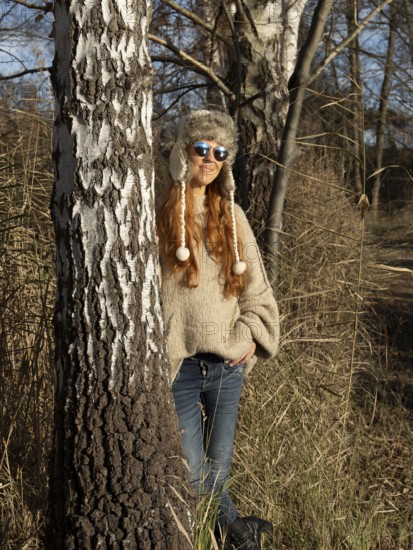 Slender pretty woman with fur hat and sweater, red hair and warm winter colors between birch forest and reeds