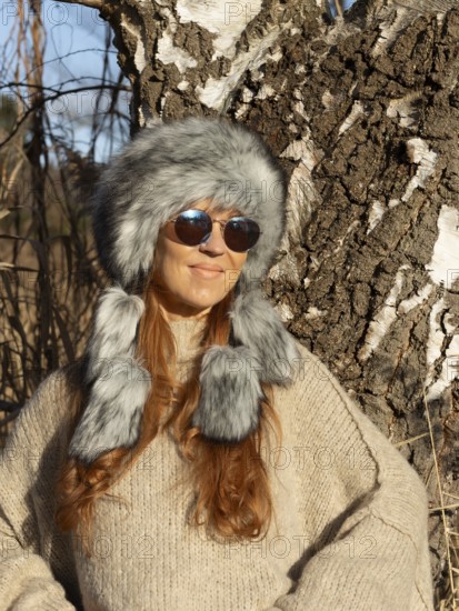 Slender pretty woman with Mongol hat, red hair and warm winter colors between birch forest and reeds