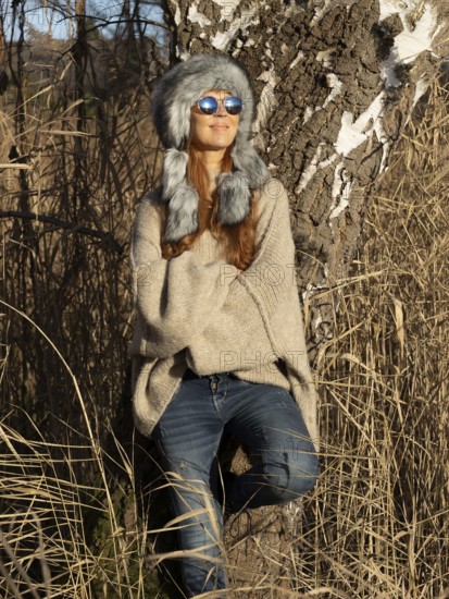 Slim woman with fur hat and sunglasses, red hair and warm winter colors between birch forest and reeds