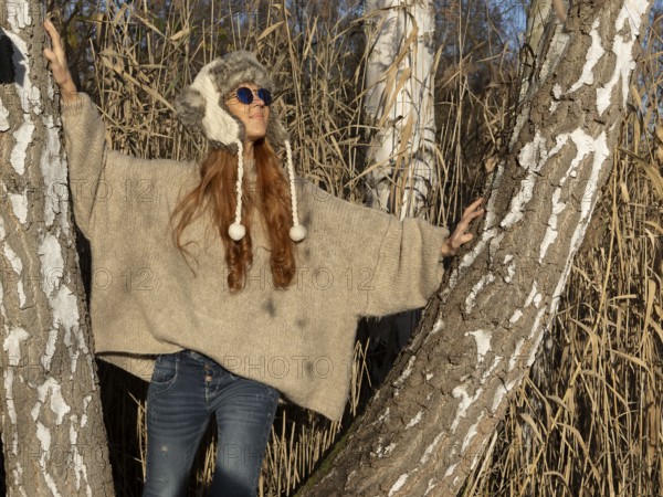Slender pretty woman with fur hat and sweater, red hair and warm winter colors between birch forest and reeds