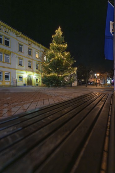 A brightly lit Christmas tree next to a building and a wooden bench, Aidlingen, Böblingen district, Germany