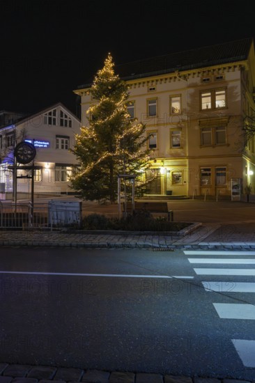An illuminated Christmas tree in front of a building and a pedestrian crossing at night, Aidlingen, Böblingen district, Germany