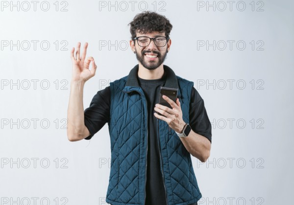 Smiling handsome man holding phone and gesturing OK, isolated. Attractive man in glasses holding phone gesturing OK