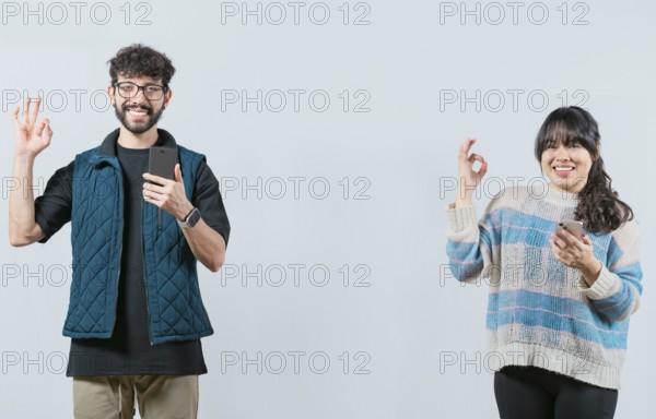 Smiling couple holding a cell phone and gesturing OK, isolated. Happy young couple holding phone gesturing OK