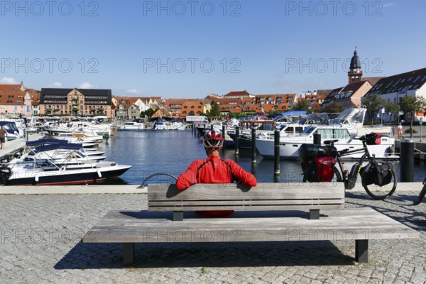 Cyclist with helmet, sitting on a wooden bench, e-bike with luggage bags, bike tour, boats in the city harbor, Waren an der MÃ¼ritz, Mecklenburg Lake District, Mecklenburg-Western Pomerania, Germany