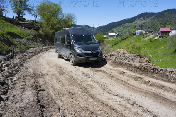 A van drives down a muddy road surrounded by green hills and individual houses, campers on the way to Goderdzi Pass, Ajara region, Adjara, Autonomous Republic, Georgia