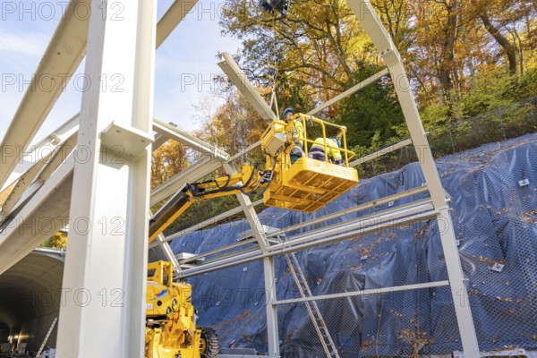 Workers in a yellow lift work on a raised metal structure in front of a forest, Hermann Hesse Railway bat tunnel construction site, Calw, Black Forest, Germany