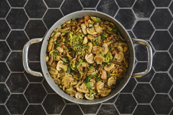 Freshly cooked mushrooms and onions are sautÃ©ed in a large skillet. Green leek add flavor. The meal is prepared in a stylish kitchen with a hexagon tile backdrop during lunch time