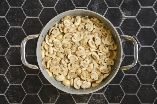 Sliced mushrooms are being fried in a large pan, giving off a savoury aroma. The scene is set on a stylish black hexagonal countertop