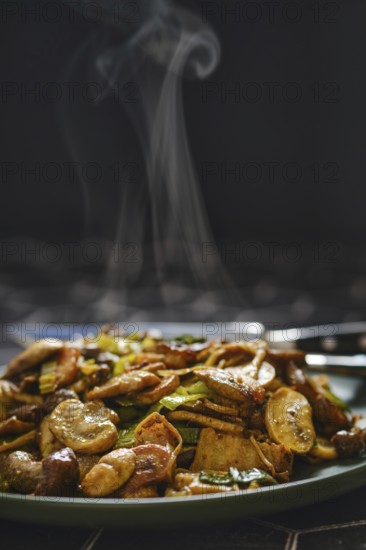 A colorful mix of mushrooms and vegetables is stir-fried to perfection, filling the air with delicious aromas. The dish steams gently as it is presented on a dark-colored plate