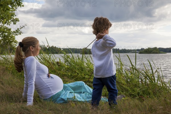 Pregnant woman with little boy at the lake, in grass, child playing with sticks