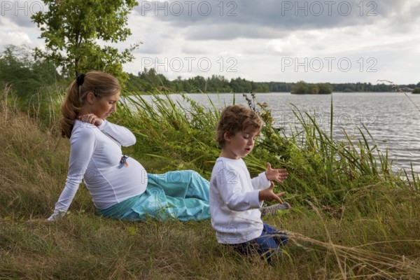 Pregnant woman thinking in grass with little boy at lake, child playing with hands