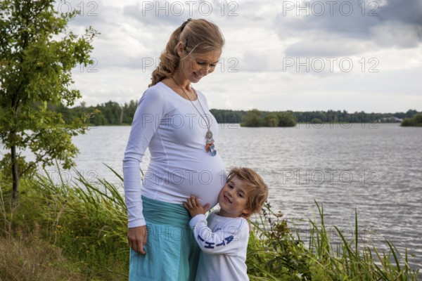 Pregnant woman happy with little boy at the lake, child listening to mom's belly