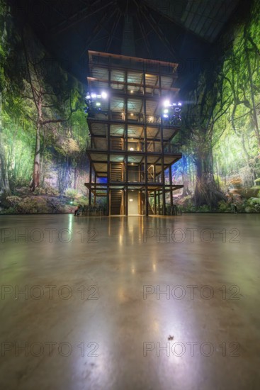 A wooden tower stands in front of a green illuminated artificial jungle backdrop, Amazonia exhibition, Gasometer, Pforzheim, Germany