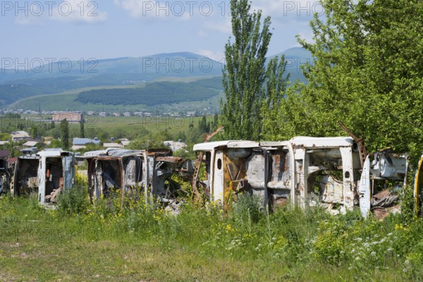 Row of rusty cars in a meadow with green trees and mountains, scrap cars serving as fencing near Zovaber, Kotayk province, Armenia