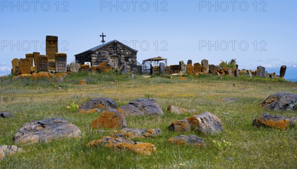 Small chapel surrounded by old tombstones on a hilly meadow under a blue sky, khachkar at the chapel of the Holy Cross and cemetery near Noratus, Sarukhan, Lake Sevan, Gegharkunik province, Armenia