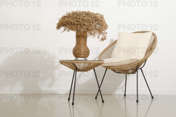 A modern space features a rattan chair with a soft pillow beside a small table. A woven vase filled with dried flowers adds a touch of nature. The minimalist design highlights calm and warmth