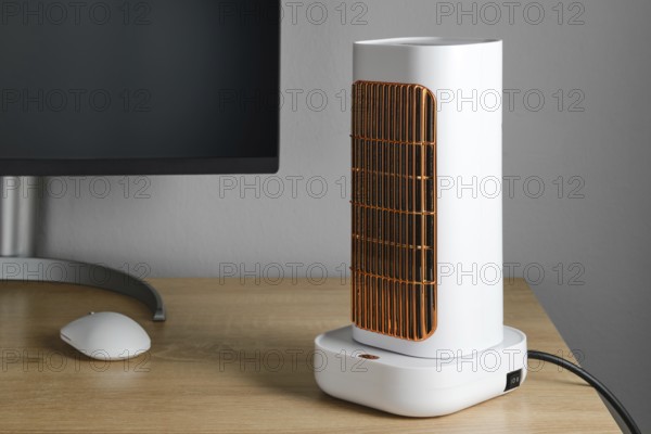 A sleek electric heater stands on a wooden desk next to a computer monitor. The heater features a unique design with a white body and an orange grill, providing warmth in a cozy workspace setting