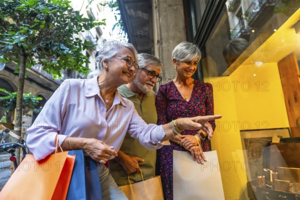 Three happy senior friends, two women and one man, standing on a city street, carrying shopping bags and pointing at items in a store window, enjoying their leisure time together
