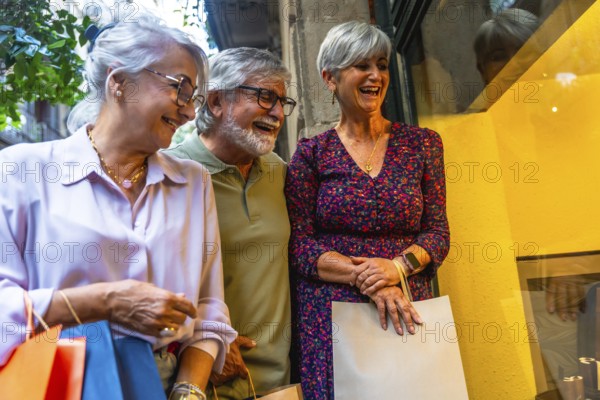 Senior friends laughing together while window shopping on a city street, carrying multiple shopping bags and enjoying their leisure time during retirement