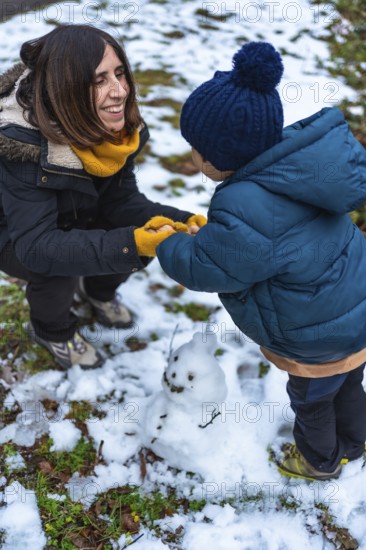 Happy mother and young son build a small snowman outdoors, laughing and bonding in warm parkas, beanies and scarves while enjoying playful winter fun and cozy family moments