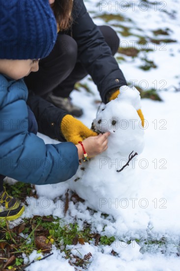 Young boy and mother collaborating to build a small snowman, carefully adding details like eyes and mouth with natural elements in a snowy outdoor setting during winter