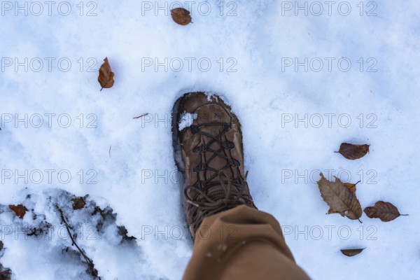 Hiker's brown boot and light pants stepping through fresh white snow scattered with dry brown leaves and twigs, close up top view conveying crisp winter trail solitude and exploration