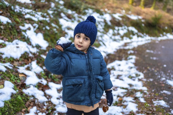 Young boy in warm parka and hat holding a snowball, smiling while playing in a snowy forest clearing, enjoying winter outdoor fun and carefree childhood adventure
