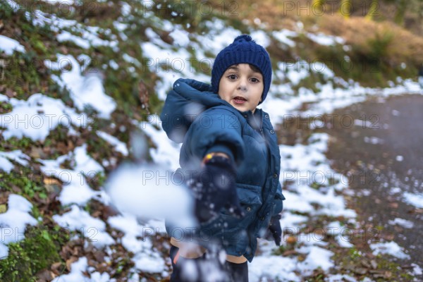 Young boy wearing a blue knitted hat and warm winter jacket, actively playing and throwing a snowball directly at the viewer, enjoying the fresh snow in a natural forest setting