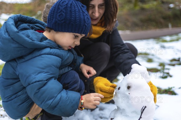 Mother and young son crouch in fresh snow, building and adding details to a tiny snowman while bundled in hats, jackets and bright yellow gloves, sharing a joyful winter moment