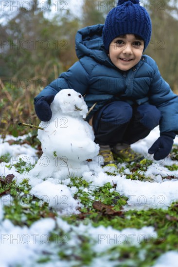 Smiling young boy crouching outdoors, happily creating a small snowman on a patch of grass lightly covered with fresh winter snow, enjoying childhood fun and creativity