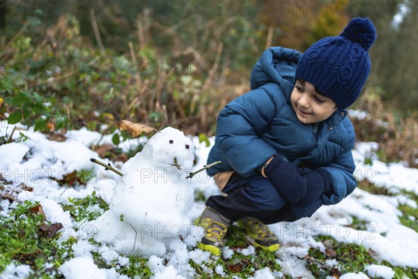 Young boy kneeling on snowy ground, wearing a warm coat, hat, and gloves, observing a freshly made small snowman with a happy expression during a cold winter day