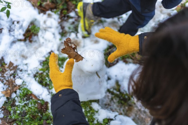 Child wearing yellow wool gloves decorates a small snowman with a brown leaf in a snowy forest, close up top view showing playful winter creativity and outdoor fun