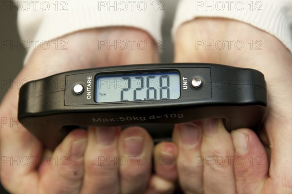 Woman holding hand scale for weight of suitcase travel bag flight luggage checks luggage weight for excess luggage in front of flight travel, scale shows 22.68 kg kilos kilograms, international