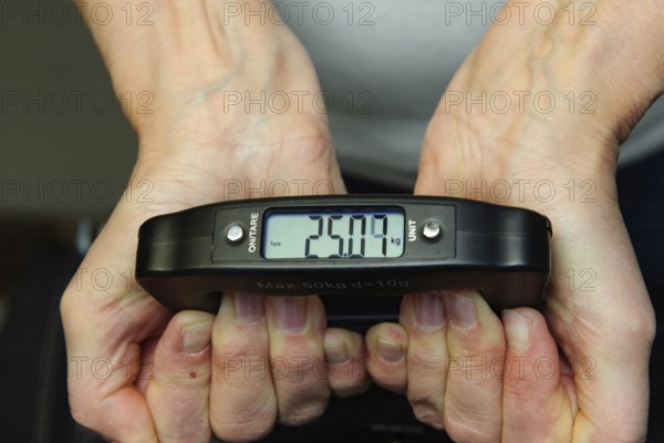 Woman holding hand scale for weight of suitcase travel bag flight luggage checks luggage weight for excess luggage in front of flight travel, scale shows 25.09 kg kilos kilograms, international
