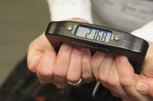 Woman holding hand scale for weight of suitcase travel bag flight luggage checks luggage weight for excess luggage in front of flight travel, scale shows 21.68 kg kilos kilograms, international