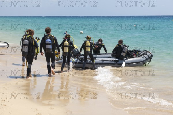 Recreational divers walk across beach to inflatable boat in diving suits with scuba tanks on their backs for boat trip trip to dive site off the coast, East Atlantic, Jandia Peninsula, Fuerteventura, Canary Islands, Spain