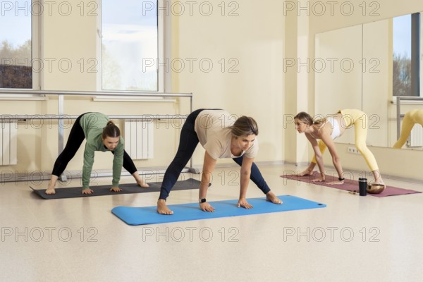 In a spacious yoga studio, three women are engaged in a group class. They are performing various poses on colorful mats, with sunlight filtering through the large windows