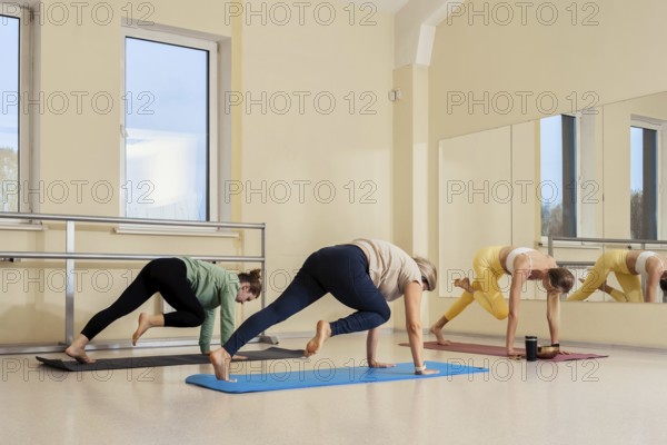 Three women engage in a fitness session in a sunny studio. They do yoga poses on colorful mats with a large mirror reflecting their focused expressions and movements