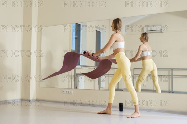 A woman prepares for a yoga session in a bright studio, unfolding her mat on the smooth floor. She wears a sports bra and leggings, ready for a morning workout. The atmosphere is calm and inviting