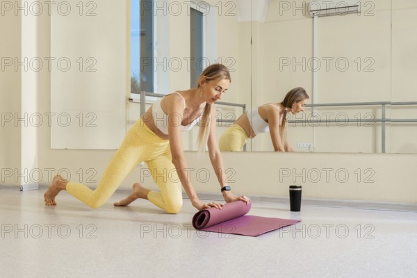 A woman in comfortable activewear rolls out her yoga mat in a well-lit studio. She is focused and preparing for her practice, with a reusable cup nearby. The mirror reflects her calm demeanor