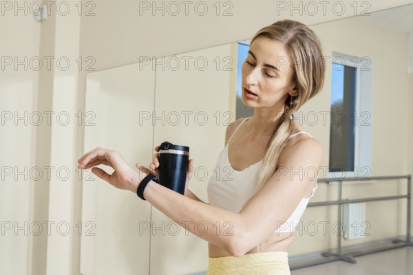 A woman in a fitness studio checks her smartwatch while holding a black drink container. She wears a white workout top and appears focused, likely monitoring her activity or workout progress