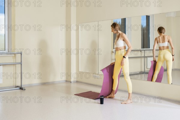 In a well-lit studio with large mirrors, a woman stands barefoot holding a yoga mat. She is dressed in a sports top and leggings, ready to start her yoga practice after setting up her space