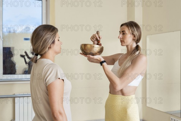 Two women engage in a sound healing session in a light-filled studio. One woman gently strikes a singing bowl while the other stands calmly, absorbing the soothing vibrations