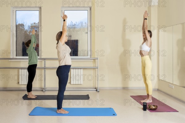 Three women are engaged in a yoga session in a spacious studio. They stretch with arms raised, promoting relaxation and fitness. Each woman uses a different colored mat