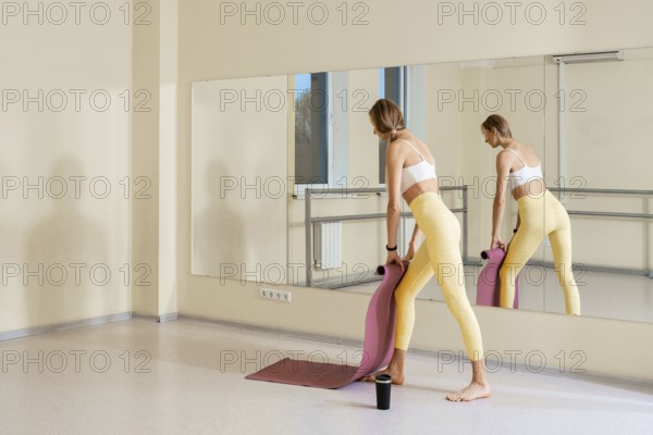 A woman in fitted yoga attire is standing in a yoga studio, preparing to practice. She stretches and uses a yoga mat, with natural light coming through large windows, creating a serene atmosphere