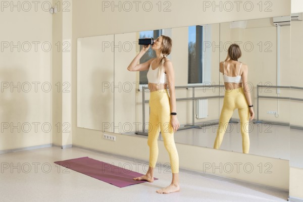 In a bright yoga studio, a woman in yellow leggings and a sports bra takes a sip from a water bottle. She stands on a mat, enjoying her practice with a mirror reflecting her focus and calmness